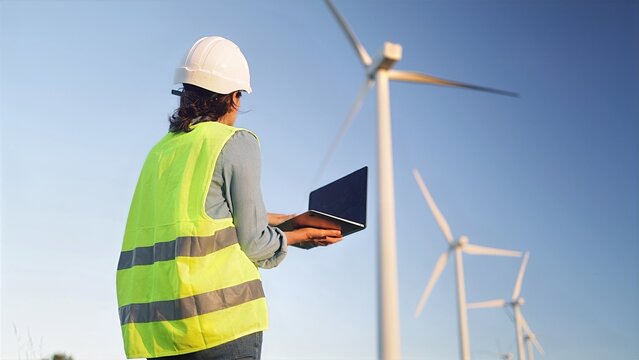 Female engineer wearing a hard hat and safety vest, looking at wind turbines while holding a laptop, overseeing clean energy production and sustainable development