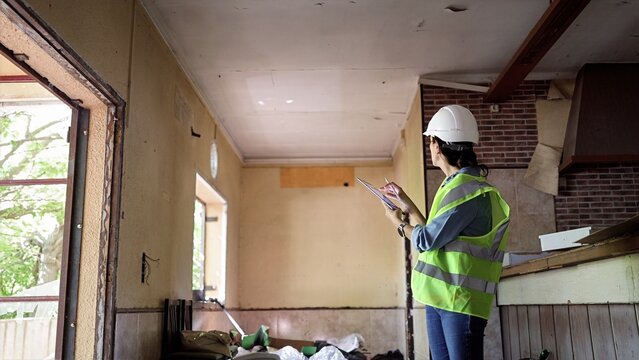 Female surveyor in hard hat and safety vest inspects and documents structural damage inside an abandoned house using a digital tablet for renovation or demolition planning and reporting