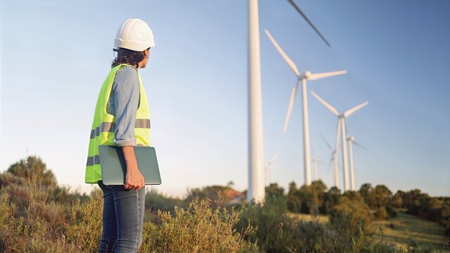 Female engineer in safety vest and hard hat holding a laptop, standing at a wind farm, and looking at the large wind turbines generating clean, renewable energy