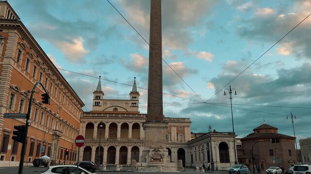 The majestic Lateran Obelisk stands tall in Piazza di San Giovanni in Laterano, Rome, Italy, with traffic passing by under a dramatic, colorful cloudy sky during late afternoon.

