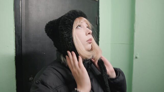 Young white woman in green hall standing in black puffer and knit hat, eyes lowered and adjusting hood, muted light, urban apartment entry, mood of isolation and introspection, implied student