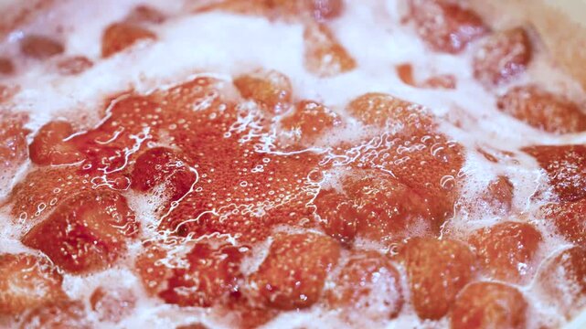 Close-up  fresh strawberries boiling in a saucepan while being cooked into homemade strawberry syrup. Juicy strawberry pieces simmer in bubbling sugar syrup, creating a rich red texture with steam