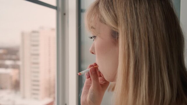 Young woman pauses to light her cigarette. Young lady stands by window lighting cigarette over cityscape. Young woman takes moment to light cigarette as snowy rooftops appear behind her