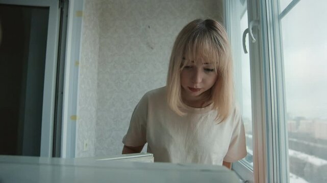 Young white woman at kitchen window, pensive domestic portrait in soft morning light, looking down at empty counter, sheer curtain and textured wallpaper in background, understated neutral palette,