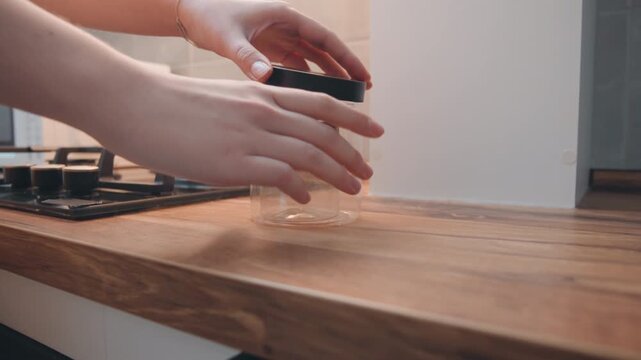 Hands sealing spice jar on counter, closeup beside gas stove, precise cap placement on empty glass container, wooden countertop texture, warm natural light, tidy domestic prep routine, focused kitchen
