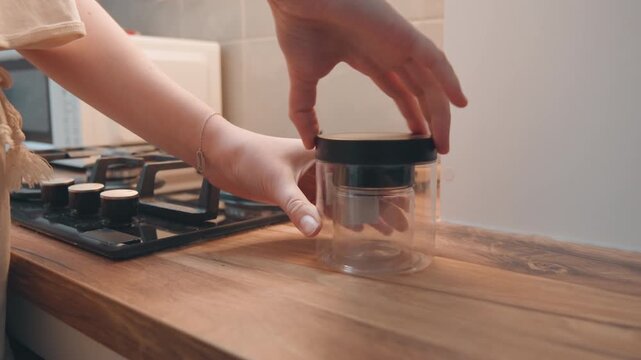 Completing kitchen storage with lid placement. Hands sealing glass jar on warm kitchen surface after cooking. Person securely covers glass container with dark lid on hot countertop