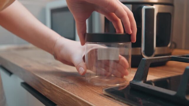 Empty glass jar on wooden counter, hand places black lid beside stove and microwave, warm natural light, tidy modern kitchen, prepping container for ingredient storage and organization