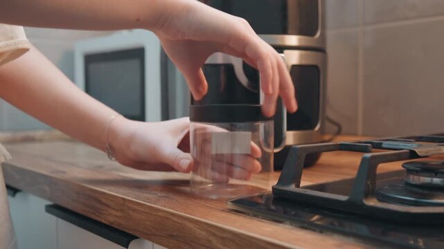 Clear glass jar and grinder lid on counter, white food blogger aligns lid by gas stove and air fryer, precise wrist motion and closeup detail show calm cooking prep and tool assembly.