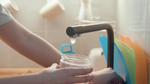 Faucet filling jar with water, soft morning light home cook holds clear jar beneath matte black tap, colorful cutting boards in background, steady pour for ingredient storage and meal prep