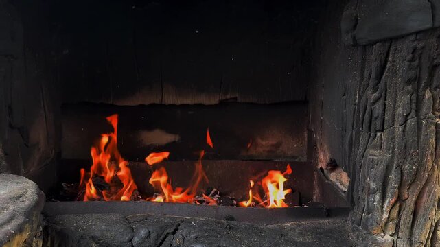 Close-up of orange flames and glowing embers inside a soot-blackened masonry fireplace, charred firebox walls and stone surround in low light, cozy indoor scene with warm contrast and copy space