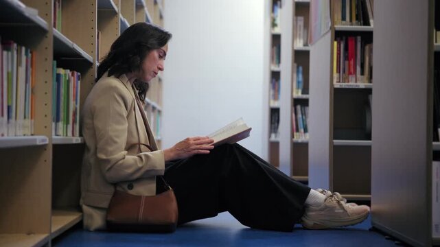 A woman sits on the floor between bookshelves in a library, engrossed in reading a book. She flips through the pages while studying.