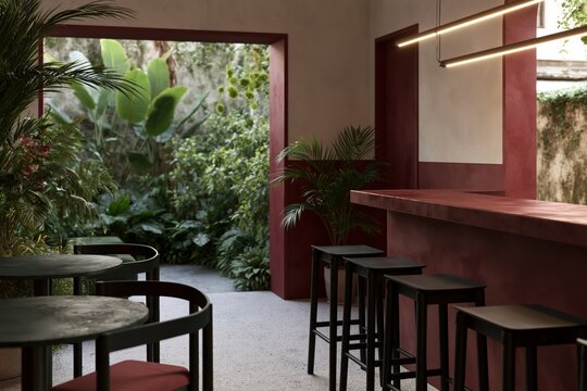 Chic Cafe Oasis: A stylish, minimalist cafe featuring a striking red bar with stools and circular tables, opens to a lush, green courtyard garden, creating a serene urban retreat.