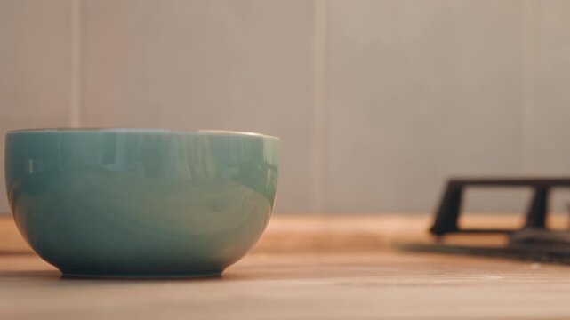 Teal bowl and single egg staged on wooden countertop near gas stove, soft morning light, hand reaches for egg, home baker preparing breakfast, calm cozy atmosphere, ceramic bowl and simple mise en