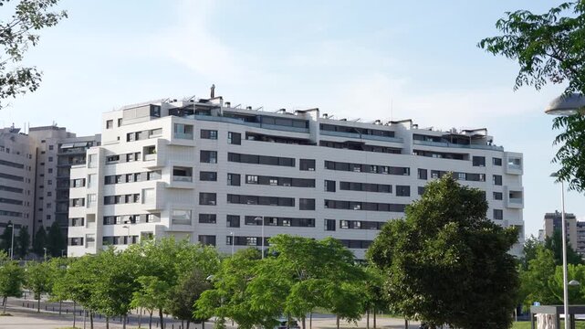 Static wide shot of a modern residential apartment building surrounded by trees and urban green spaces in Valdebebas, Madrid, Spain. Contemporary housing development and sustainable urban living conce