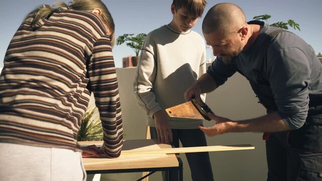 Father and teenage children working together on sunny terrace, sawing wooden plank as part of DIY home project, showing teamwork, cooperation, and family bonding.