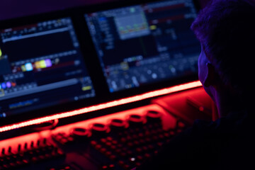 Fototapeta premium Individual with short hair working at dual computer monitors displaying colorful graphics and data, illuminated by red LED lights in a darkened workspace