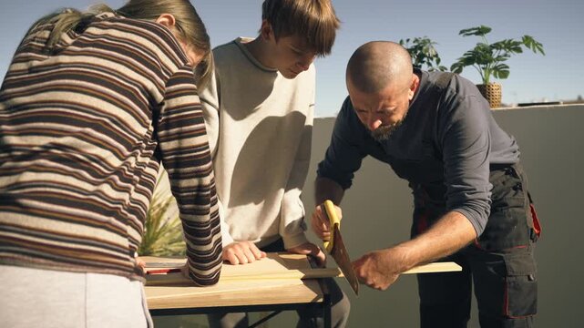 Father teaching his teenage son and daughter woodworking skills while they saw wooden plank together on outdoor terrace during practical DIY training.