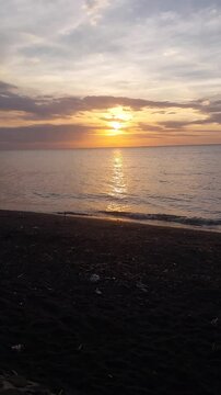 Beautiful golden sunset over a black sand beach with calm sea water and colorful evening sky. Peaceful coastal scenery with warm sunlight reflection, located at Singaraja, Bali, Indonesia.