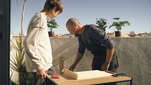 Father showing teenage son how to measure and mark wooden board with ruler and tape measure before cutting, demonstrating accurate planning and construction skills.
