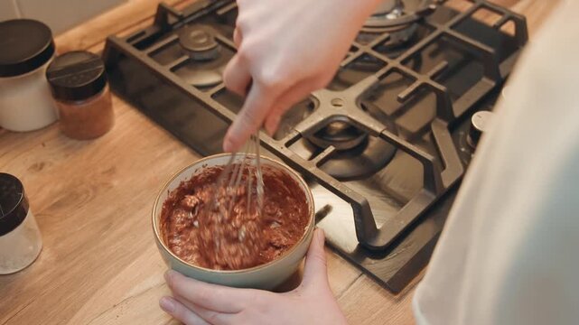 White hands whisking chocolate batter bowl, closeup view of homemade batter being mixed with whisk on wooden countertop near stove, jars of sugar and spice, warm natural light, cozy baking scene