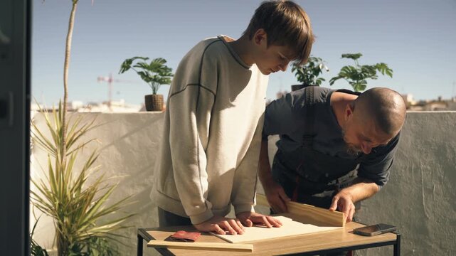Father helping teenage son prepare wooden board for DIY home project, showing how to measure and cut materials during family hands-on construction activity.