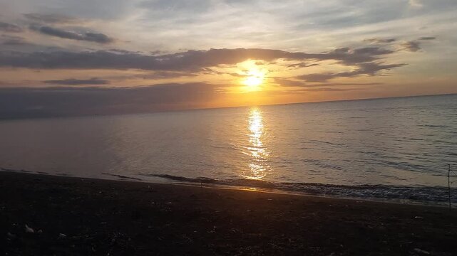 Beautiful golden sunset over a black sand beach with calm sea water and colorful evening sky. Peaceful coastal scenery with warm sunlight reflection, located at Singaraja, Bali, Indonesia.
