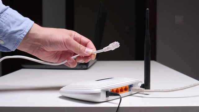 Person's hand connecting a white ethernet patch cord to a wireless internet router on a white table, establishing network connectivity with a laptop in the background