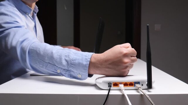 Angry man in a blue shirt slams his fist on a wireless router after struggling with slow internet and network connection problems at home, symbolizing digital frustration