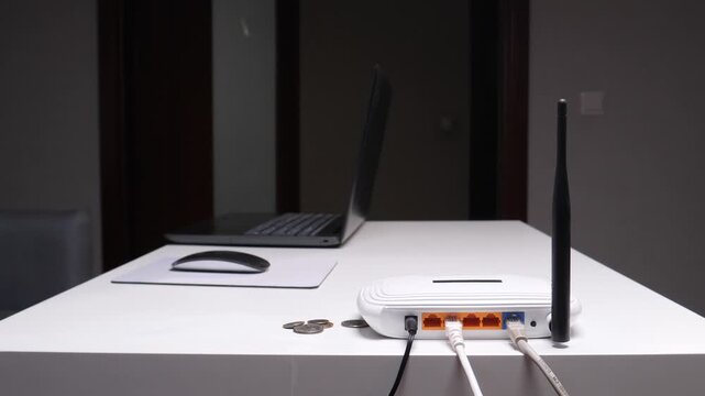 Persons hand dropping coins onto a table next to a wireless router and laptop, symbolizing the recurring expense and financial cost of internet services
