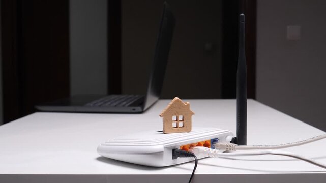 Wooden house model sitting on a white wireless router with connected cables, representing home internet connectivity and smart solutions for modern living, with a laptop in the background