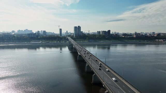 Aerial view of a long urban bridge spanning a wide river, carrying steady traffic toward a modern city skyline, with water reflections, high-rise towers, cranes, and greenery.