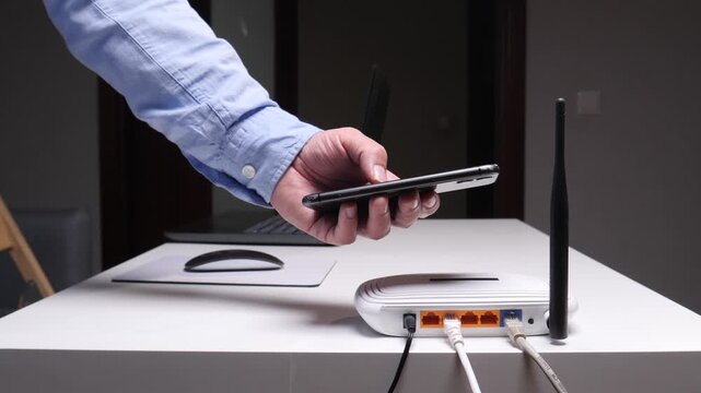 Person's hand interacting with a smartphone near a wireless internet router with connected cables on a white table, symbolizing modern connectivity, networking, and smart home technology