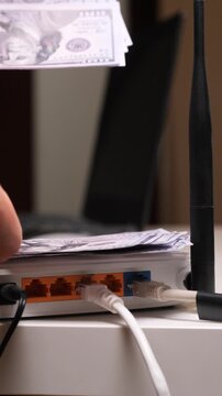 Vertical video hand holding a us dollar banknote, adding it to a stack of money on top of a connected wi fi internet broadband router, representing the concept of internet costs and expenses