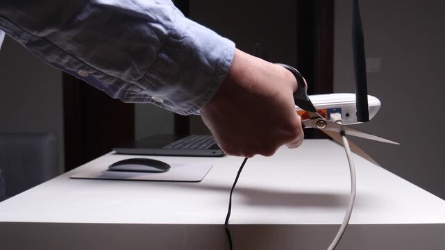 Person's hand holding open scissors, cutting a white network cable plugged into a wifi router on a white desk, symbolizing disconnection, digital detox, or internet interruption