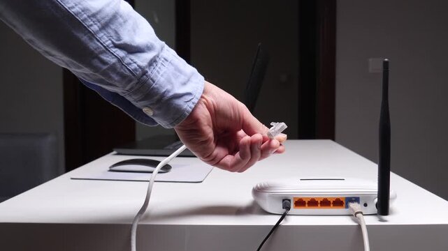Hand holding an ethernet cable as its plugged into a white wireless router on a desk, setting up a wired internet connection for home or office networking and connectivity