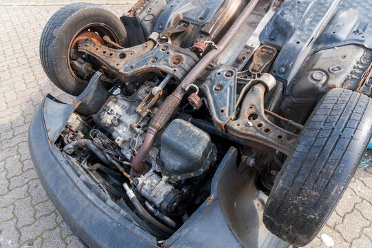 Close-up of an overturned car underbody showing wheels, exhaust, suspension, transmission, rusted frame, and exposed mechanical parts. Useful for auto repair, scrapyard, accident damage, and vehicle r