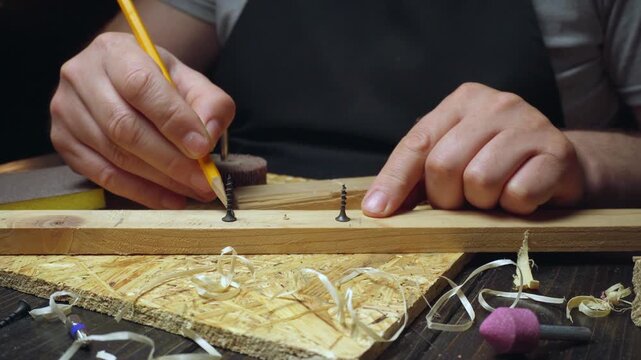 Skilled craftsman marks positions for screws on wooden board using pencil, surrounded by tools and wood shavings in a workshop setting