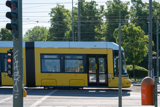 Germany, Berlin, May 25, 2026. A yellow Berlin tram is seen close up at a street intersection with red traffic lights. Overhead wires, green deciduous trees and an orange bin frame the transport scene