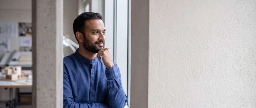 Indian businessman looking out window in an office thinking about future career success, dreaming and contemplating next strategy in modern design studio architectural office with concrete wall space.