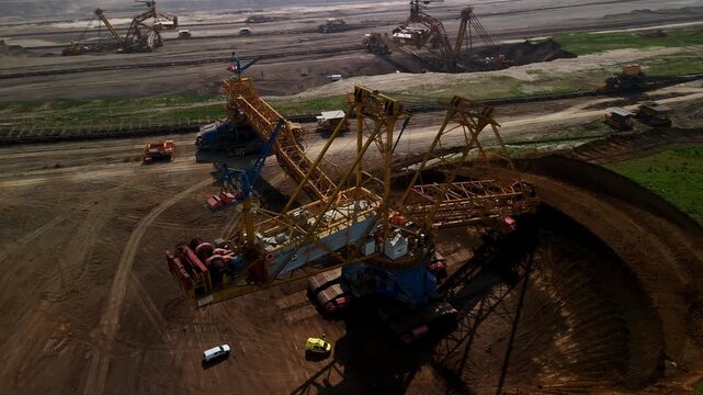 Horizontal aerial drone view of a bucket wheel excavator in the foreground with additional mining machines visible across a vast open pit quarry landscape.
