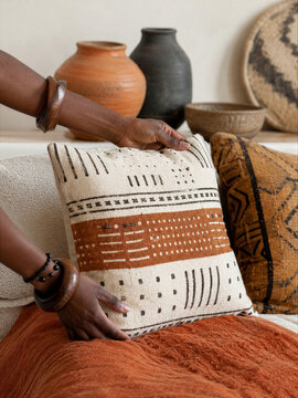 Woman hands arranging mudcloth cushion on sofa in living room. Afro bohemian interior design with ethnic textile and clay vessel. Home decoration style with natural fabric texture.