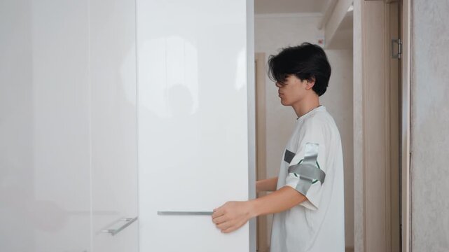 Opening glossy wardrobe doors in hallway asian teen pulls white cabinet door, inspects shelves and handles, minimalist corridor in small apartment, quiet declutter routine preparing clothes