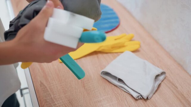 Sponge, cloth, gloves on wooden counter. White homeowner presses melamine sponge into microfiber cloth, prepping cleaning tool for kitchen surface maintenance, tidy domestic routine, closeup hands,