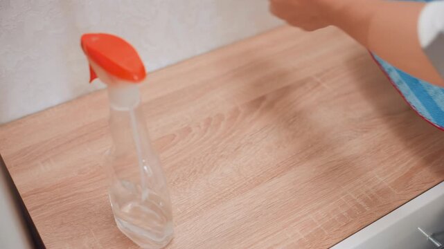 Spray bottle and mat on counter. White caretaker places trigger spray, lines up gloves and sponge, prepares gentle solution for wood surface refresh, calm domestic routine, closeup hands and tools