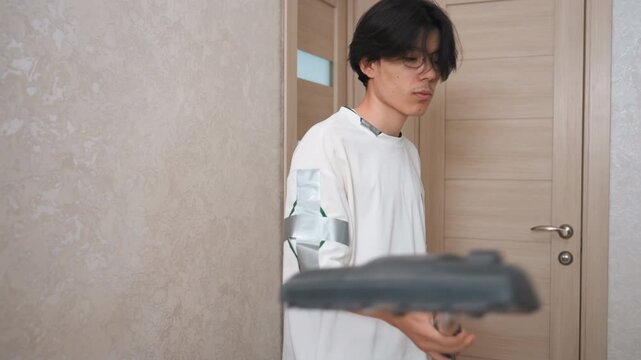 Asian man testing vacuum near door with casual pose, checking attachment and handle, modern apartment interior, wooden door and neutral wallpaper, lighthearted mood demonstrating household cleaning