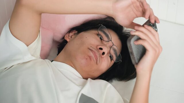 Youthful Asian professional. Person browsing phone at home. Young professional relaxing with phone in cozy bedroom. Asian young adult enjoys leisure time alone with device in comfortable home setting