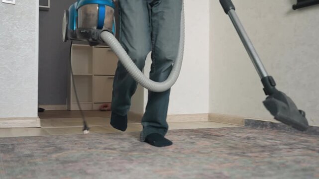 Asian man vacuuming patterned carpet closeup showing nozzle and wheels, blue canister vacuum gliding over textured pile, hose and cord in frame, focus on motion and suction, quiet efficient cleaning,