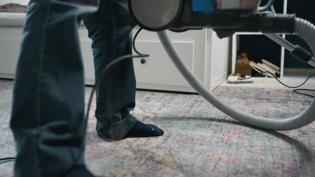 Lowangle vacuuming on living room carpet. Homeowner uses blue canister vacuum to tidy patterned rug, hand lifting unit and guiding nozzle, casual weekday chore in cozy interior with soft light