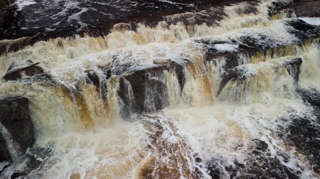 Approaching an overflowing, slow motion waterfall from above. View approaches the waterfall until above it looking down, then rises. Water is golden and turbulent. Lots of detail.