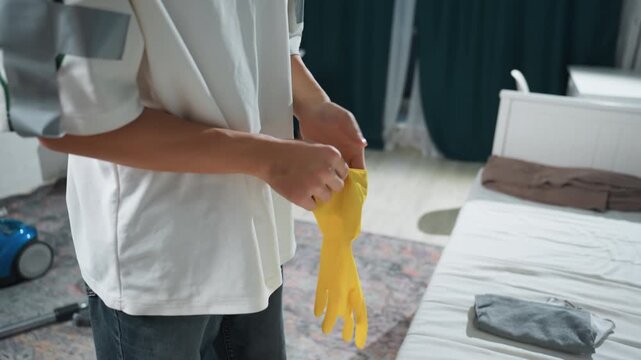 Asian man puts on yellow gloves before cleaning bedroom, homeowner preparing mattress and vacuum, protective rubber gloves, tidy modern apartment, calm morning routine, focus on hands and texture